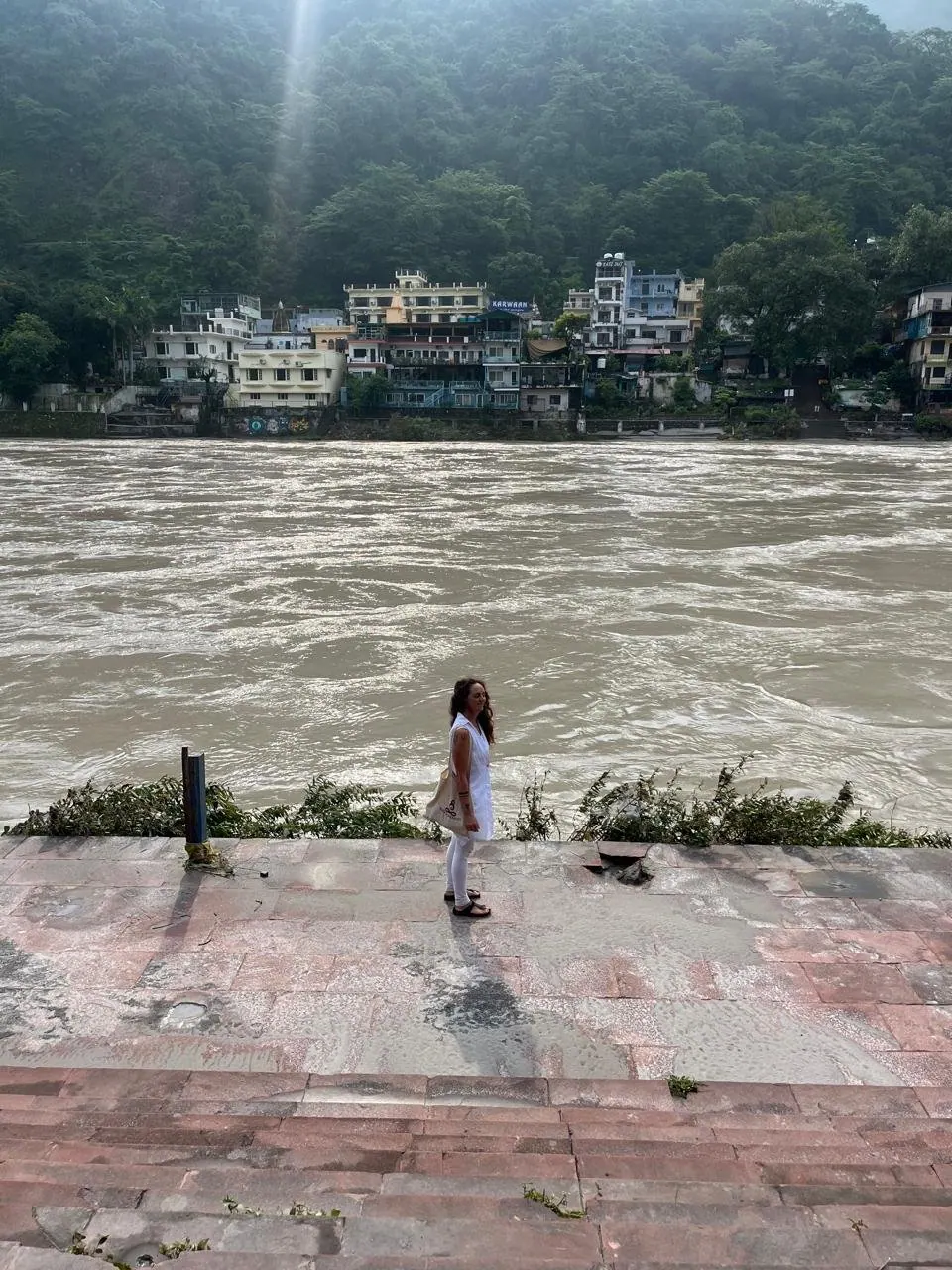 Laura in bianco sulla riva del Gange a Rishikesh, con il fiume impetuoso e le case sulla collina alle spalle