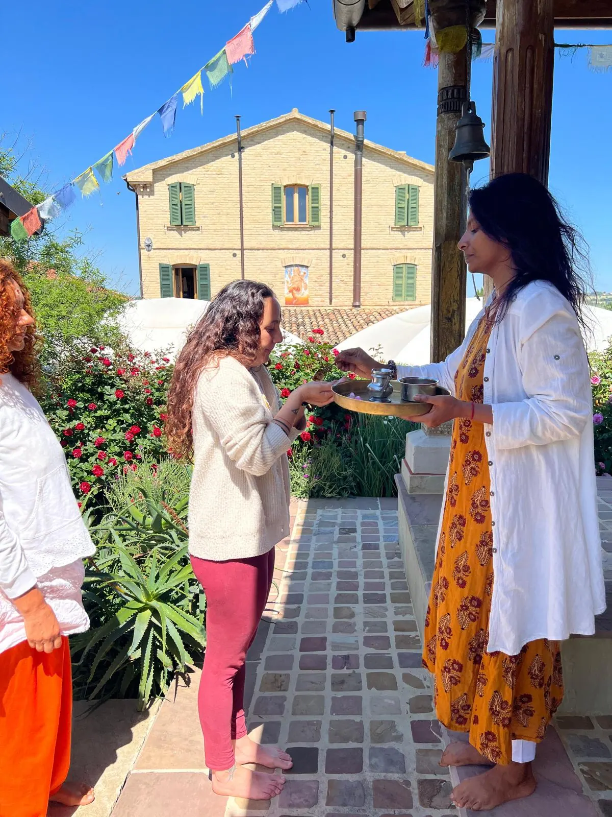 Due donne si scambiano un'offerta sotto un portico con le bandierine di preghiera, davanti a una casa gialla