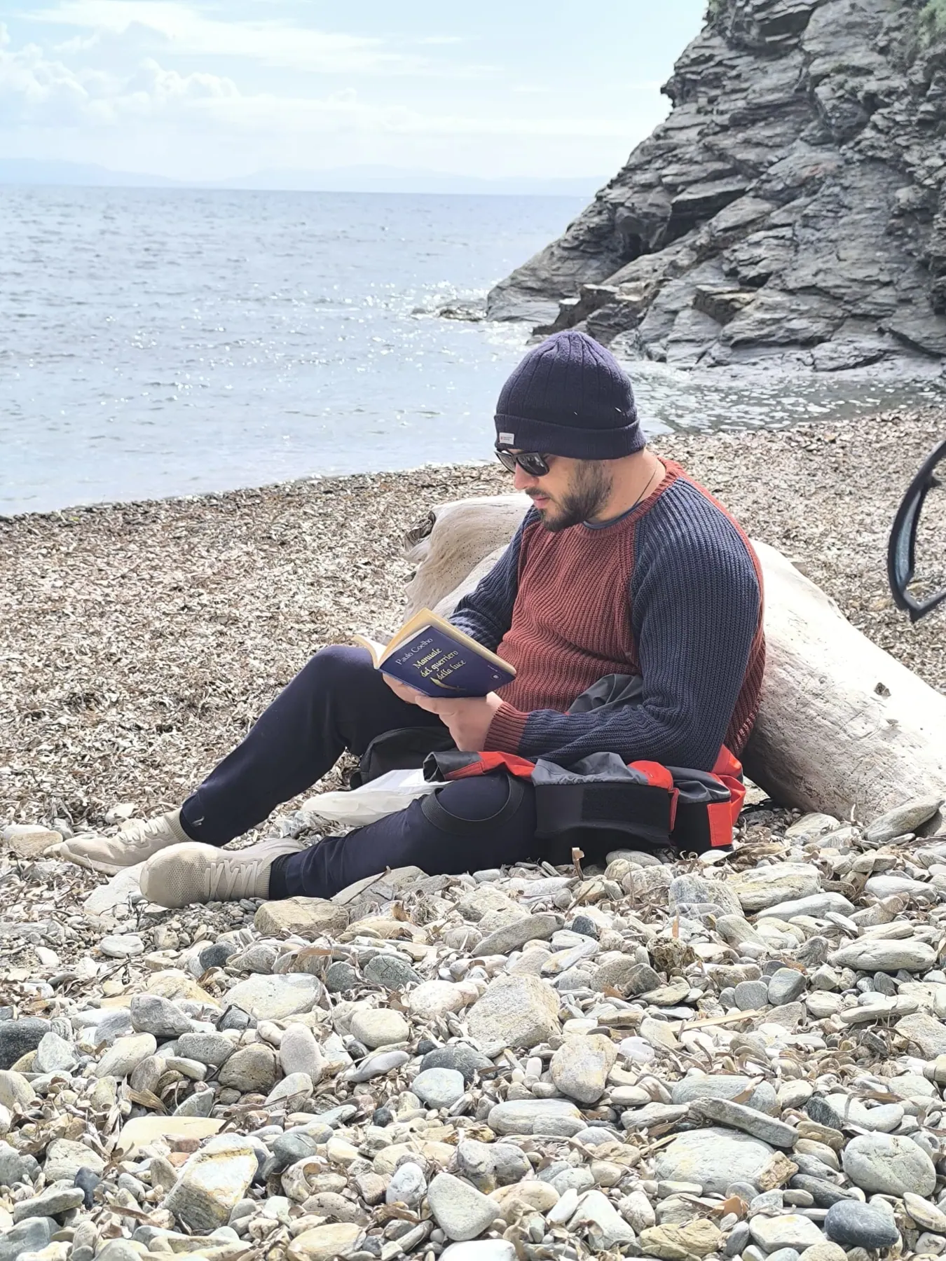 Un uomo legge seduto sulla spiaggia di ciottoli, appoggiato a un tronco, davanti al mare