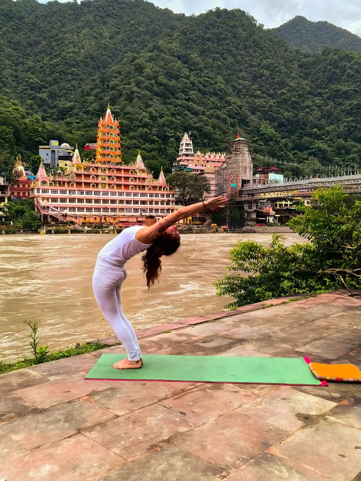 Laura in un piegamento all'indietro sul tappetino, sulla riva del Gange davanti ai templi di Rishikesh